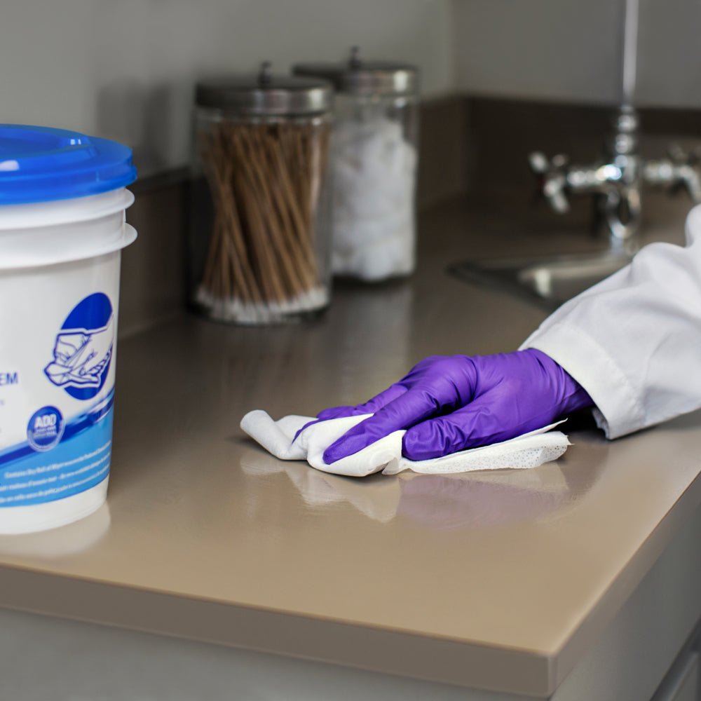 Osoba s fialovými rukavicami utiera WypAll® Wettask™ utierkami od Kimberly-Clark GmbH (6 roliek x 90 listov) béžovú pracovnú dosku blízko umývadla, zatiaľ čo v pozadí sú viditeľné poháre s vatovými tyčinkami a guľkami.