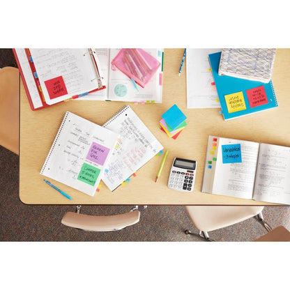 Stôl v triede, úhľadne usporiadaný s otvorenými zošitmi, učebnicami, kalkulačkou, farebnými perami, zložkami a Post-it® Notes z Energetic Collection (38mm x 51mm, 100 listov/blok), značka: 3M Deutschland GmbH; tri prázdne stoličky obklopujú stôl.
