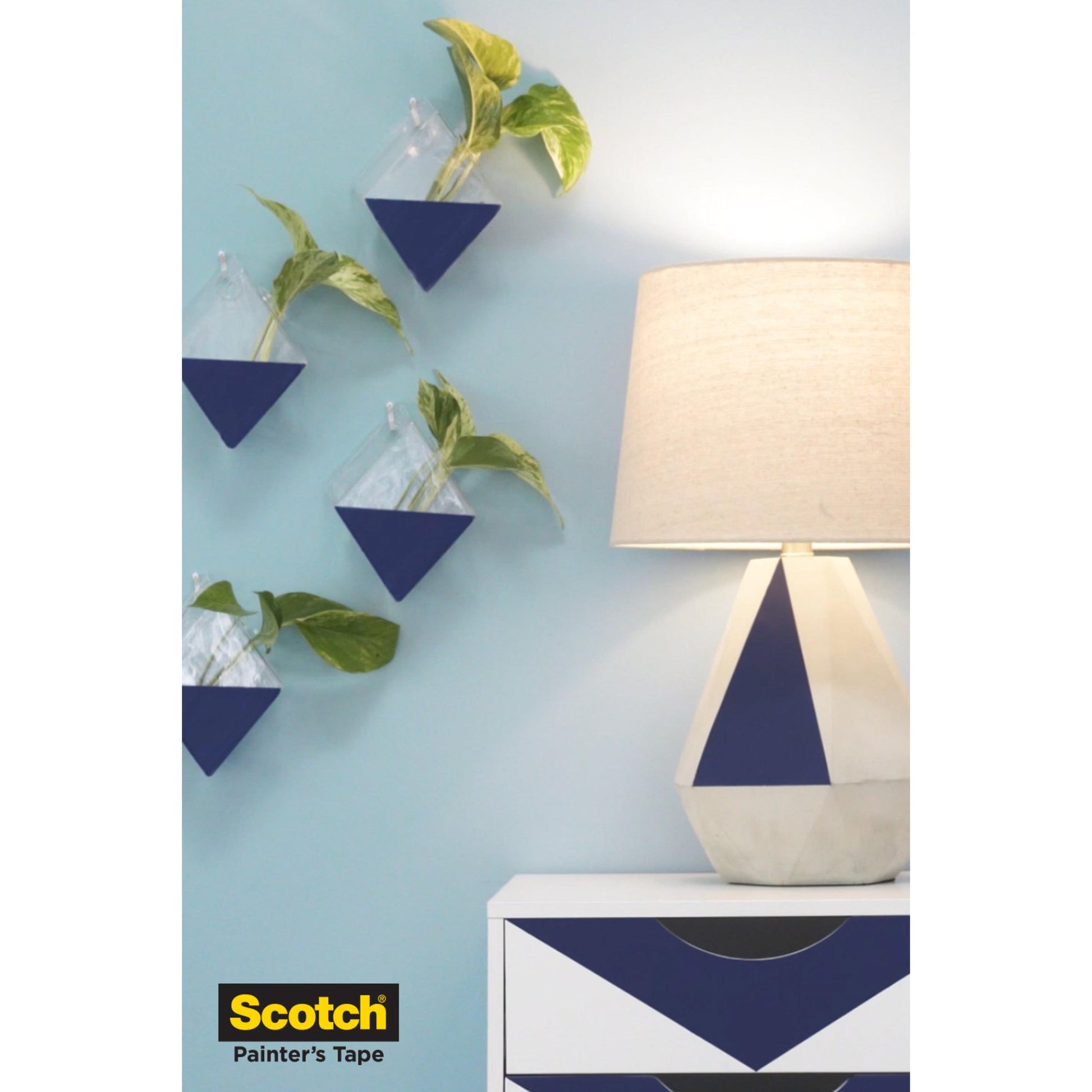 Eine weiße und blaue geometrische Tischlampe steht auf einer passenden Kommode neben einer hellblauen Wand mit drei dreieckigen Pflanzgefäßen mit Grünpflanzen. Das Scotch® Malerabdeckband für empfindliche Oberflächen by 3M Deutschland GmbH Logo befindet sich in der unteren linken Ecke.
