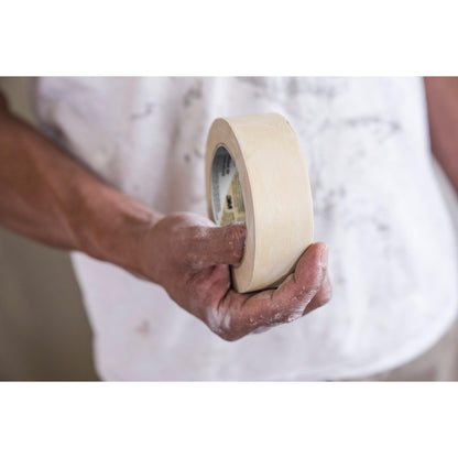 Eine Person mit farbverschmierten Händen, in einem farbverschmierten weißen Hemd, hält eine Rolle 3M Deutschland GmbH Scotch® Classic Abdeckband (36 mm x 50 m, 70% PEFC, SGSCH-PEFC-COC-110078); der Hintergrund ist unscharf.