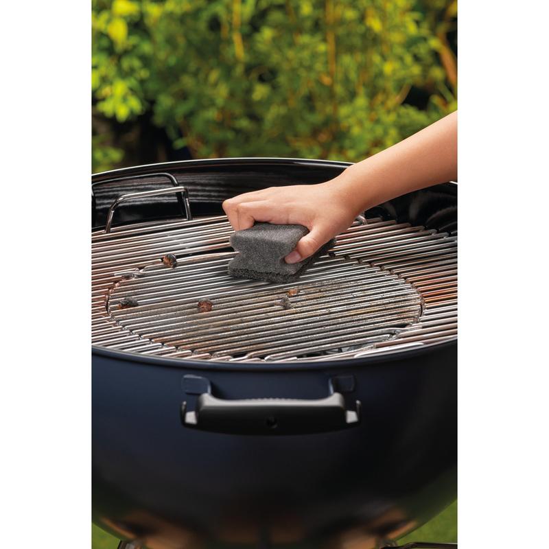 Ruka čistí kovový gril okrúhleho grilu na drevené uhlie s použitím Scotch-Brite® Grillgriffschwamm (1 kus na balenie) od 3M Deutschland GmbH. V pozadí je rozmazané zelené lístie.