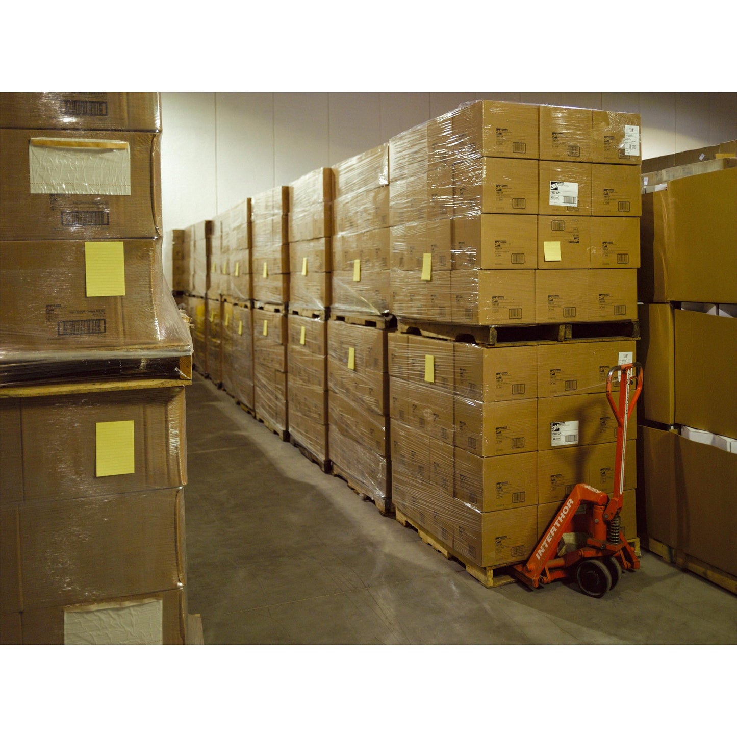 V sklade sú naskladané Post-it® Super Sticky Notes vo veľkom formáte, Ultra Žltá (101 x 152 mm, 90 listov/blok, 6 blokov), od 3M Deutschland GmbH, zabalené na paletách. Vedľa stojí červený ručný paletový vozík.