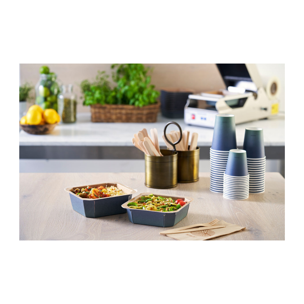 Na stole sú dve misky na odniesť, obklopené naskladanými Duni Urban Kaffeebecher PAP/PLA od Duni GmbH, dreveným príborom v držiaku a servítkami. Citróny, poháre a bylinky v pozadí prispievajú k udržateľnej atmosfére tohto priestoru na prípravu jedál.